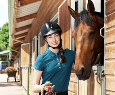 Eine lächelnde Frau mit Reithelm steht neben einem Pferd, das aus einem Stall herausschaut.