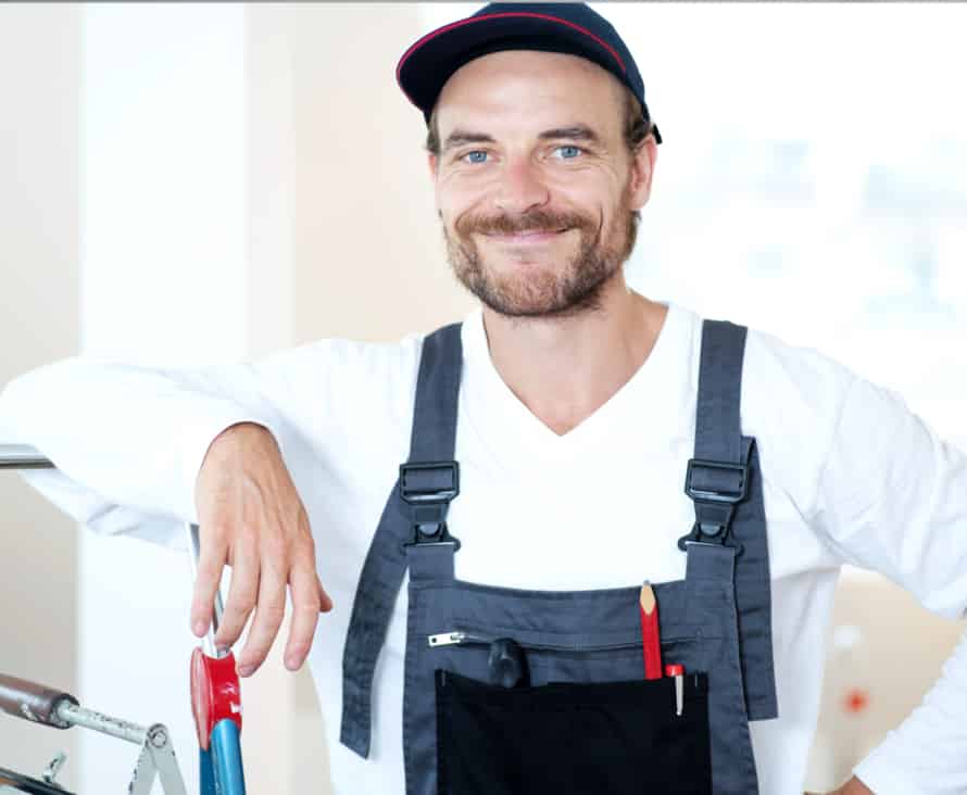 Ein lächelnder Mann mit Mütze und Overall lehnt in einer hellen Werkstattumgebung auf einem Fahrrad und hält einen Schraubenschlüssel in der Hand.