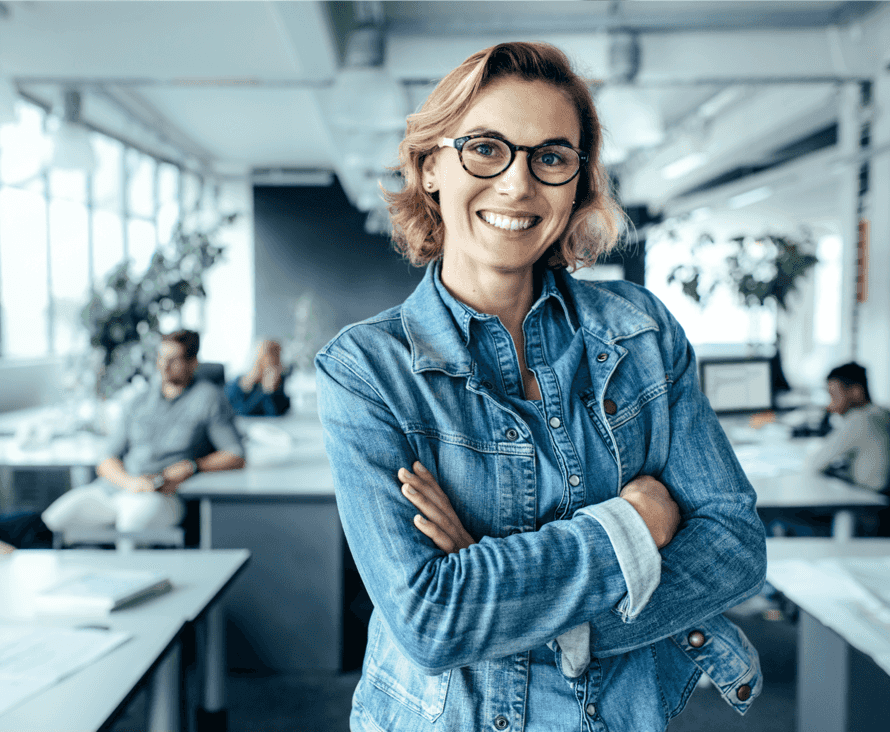 Eine selbstbewusste Frau in Jeansjacke und Brille steht mit verschränkten Armen in einer geschäftigen Büroumgebung.