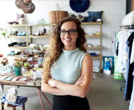 Eine fröhliche Frau mit lockigem Haar und Brille steht mit verschränkten Armen in einer Boutique voller Kleidung und Körbe.