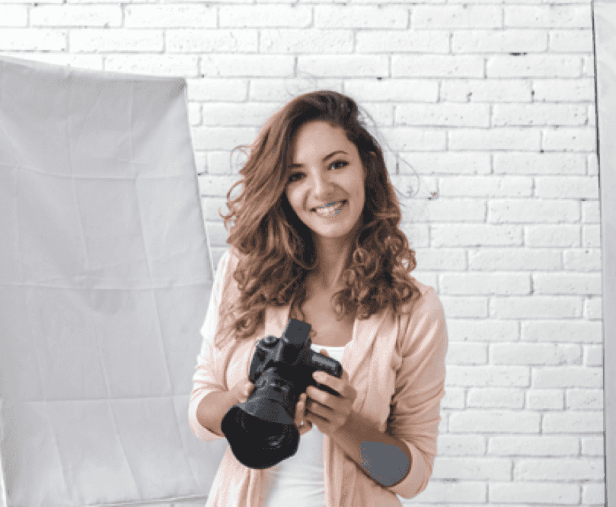 Eine lächelnde Frau mit lockigem Haar hält eine Kamera und steht vor einer weißen Backsteinwand.