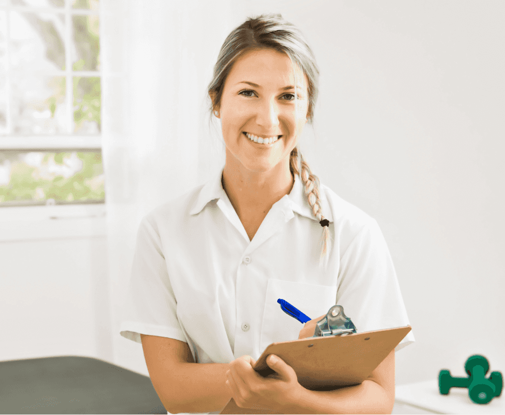 Eine lächelnde Frau in einer weißen Uniform hält ein Klemmbrett und steht in einem hellen Raum mit einem Fenster im Hintergrund.