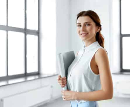 Eine berufstätige Frau hält einen Ordner, lächelt sanft und steht in einem hellen Büroraum mit großen Fenstern.