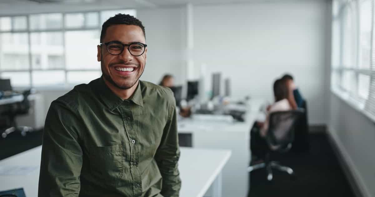 Lächelnder Mann mit Brille und grünem Hemd steht in einem modernen Büro mit Kollegen im Hintergrund.