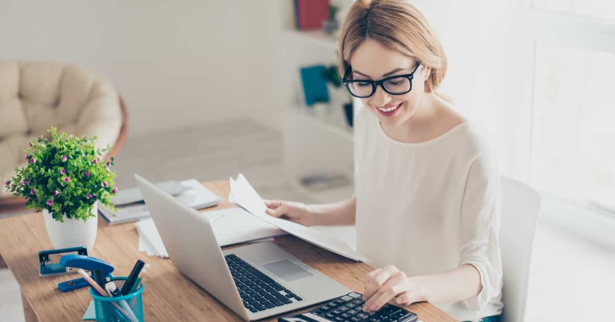 Eine Frau mit Brille lächelt und überprüft Dokumente, während sie an einem Schreibtisch mit einem Laptop und einer grünen Pflanze neben sich sitzt.