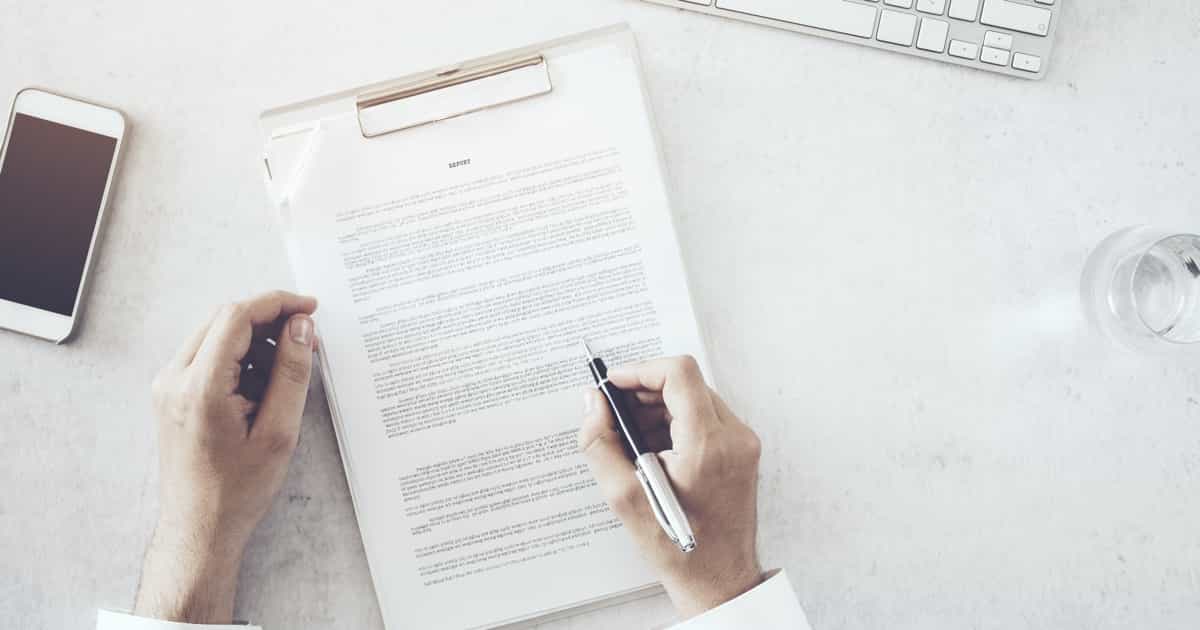 Person überprüft und unterschreibt ein Dokument an einem Schreibtisch mit einer Tastatur und einem Glas Wasser in der Nähe.