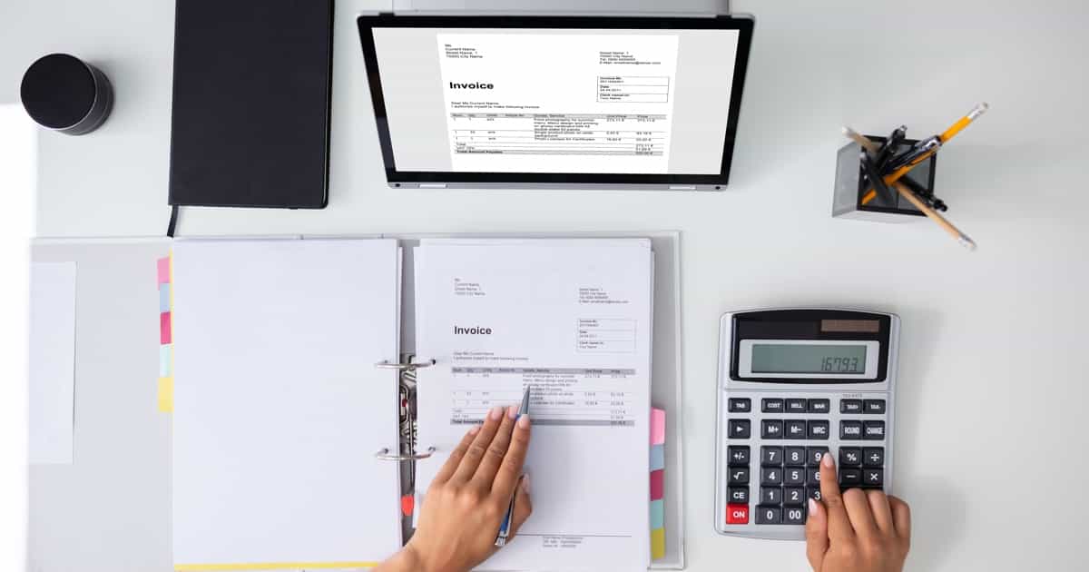 Draufsicht auf einen Arbeitsbereich mit einer Person, die einen Taschenrechner verwendet und Dokumente überprüft, darunter einen offenen Ordner und eine Rechnung auf einem Laptop-Bildschirm.