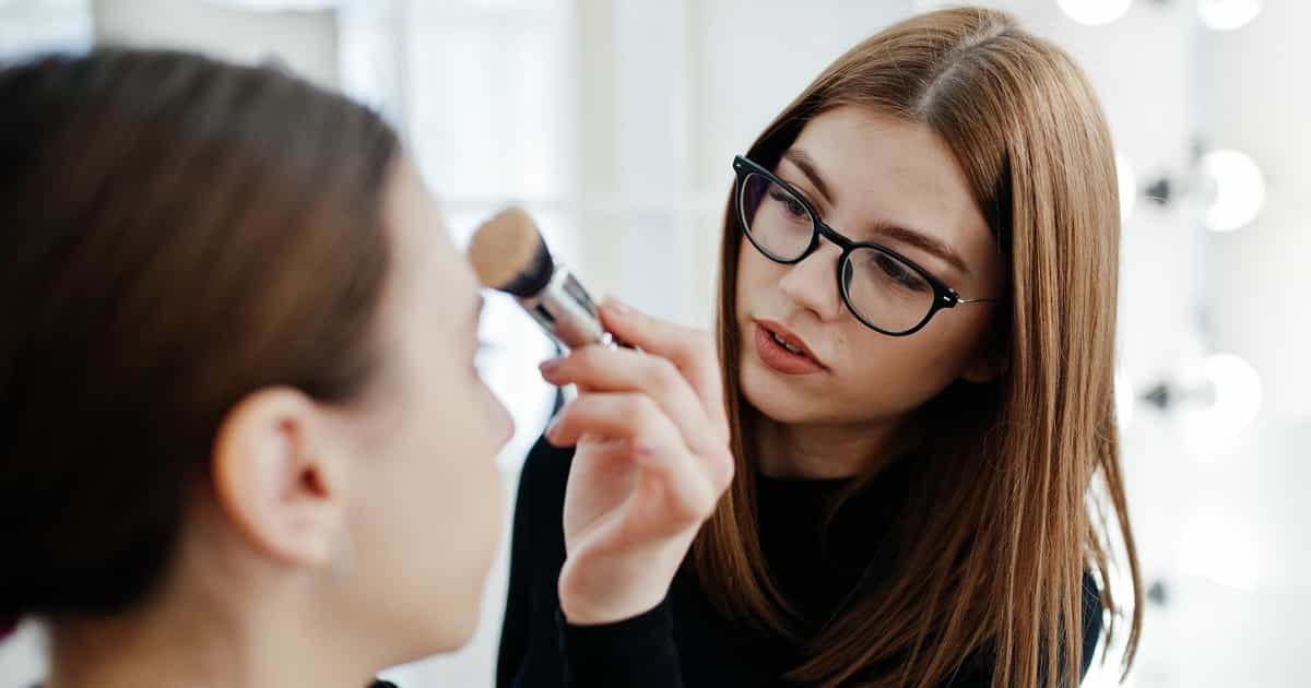 Eine Visagistin schminkt eine junge Frau mit Brille in einem gut beleuchteten Raum.