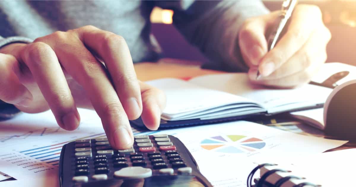 Eine Person verwendet einen Taschenrechner, während Finanzdiagramme und -dokumente auf einem Schreibtisch ausgebreitet sind, was darauf schließen lässt, dass sie mit Budgetplanung oder Buchhaltungsarbeiten beschäftigt ist.