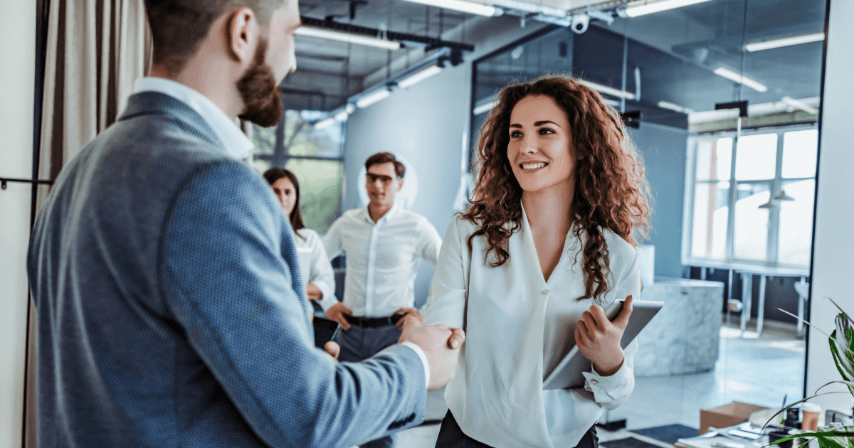 Eine Frau mit lockigem Haar schüttelt einem Mann in einem modernen Büro die Hand, im Hintergrund beobachten drei Kollegen.