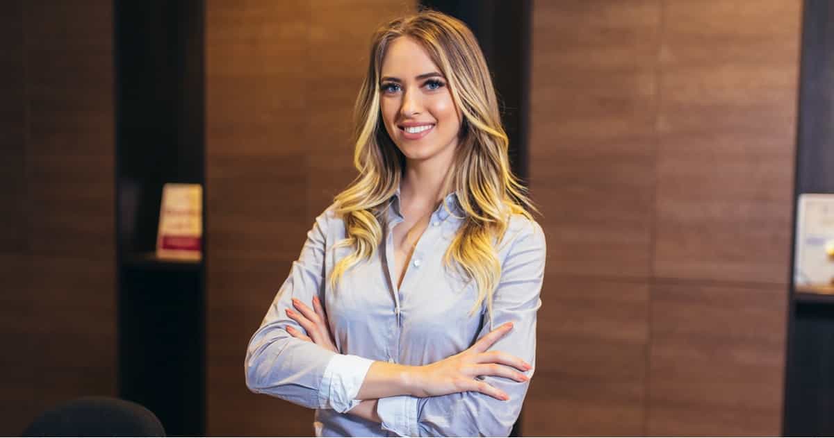 Eine selbstbewusste junge Frau im legeren Business-Outfit steht mit verschränkten Armen in einer Büroumgebung.