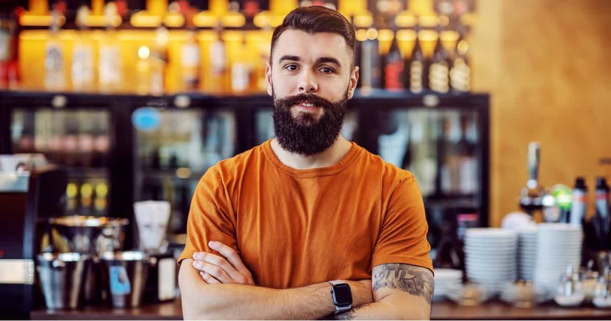 Ein bärtiger Mann in einem orangefarbenen T-Shirt steht selbstbewusst mit verschränkten Armen vor einer Bar, hinter ihm stehen Regale voller Flaschen.