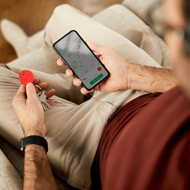 Ein auf einer Couch sitzender Mann nutzt ein Smartphone, um seine Schlüssel mit einem roten Ortungsgerät zu orten.