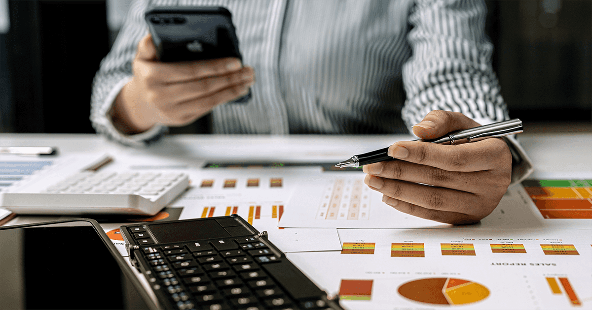 Person sitzt am Tisch mit Stift und Handy in der Hand und führt Berechnungen durch