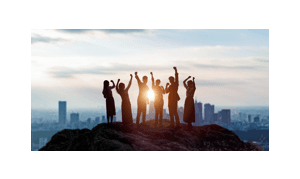 Eine Gruppe von Menschen auf einem Felsvorsprung feiert mit erhobenen Armen vor der Skyline einer Stadt bei Sonnenuntergang.