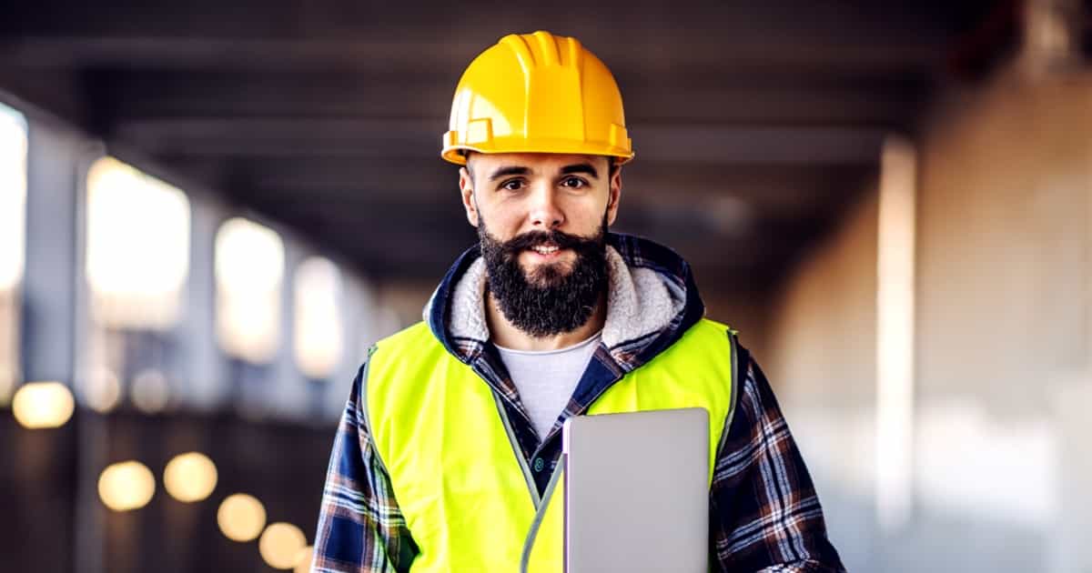 Ein Bauarbeiter mit gelbem Helm und Warnweste hält ein Tablet in der Hand und steht in einer halb geschlossenen Umgebung.