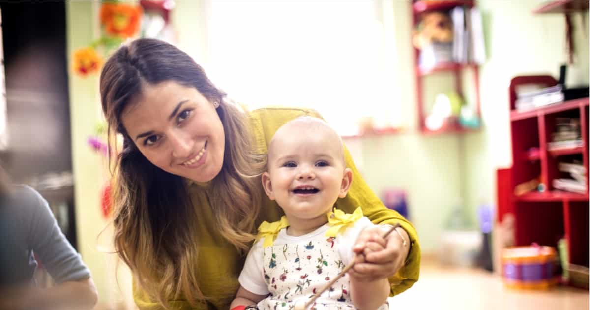 Eine lächelnde Frau hält ein glückliches Baby in einem gelben Outfit und sitzt in einem farbenfrohen Spielzimmer.
