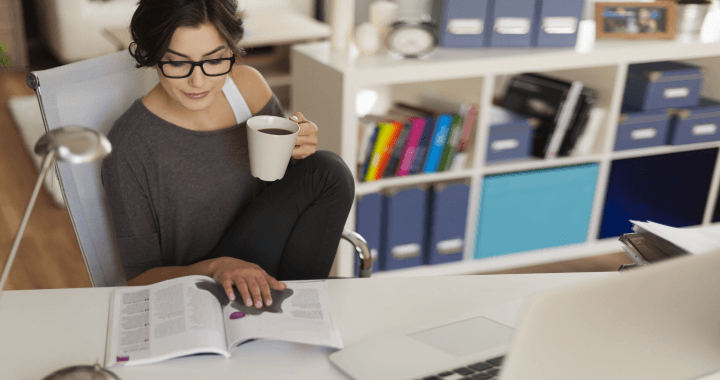 Frau, die mit gekreuzten Beinen auf einem Stuhl sitzt, ein Buch liest und eine Tasse hält, im Hintergrund ein Laptop und ein aufgeräumtes Regal.