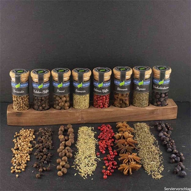 Verschiedene Gewürze in Gläsern auf einem Holzregal vor dunklem Hintergrund, davor kunstvoll arrangierte lose Gewürze.