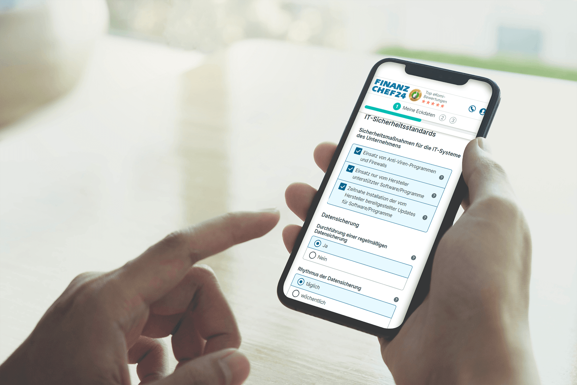 Person bedient ein Smartphone, das eine Finanzchef24-Seite mit einer Abfrage zu IT-Sicherheitsvorkehrungen beim Abschluss einer Cyberversicherung anzeigt. Es werden Fragen zur Nutzung von Antivirenprogrammen und zur Datensicherung gestellt.