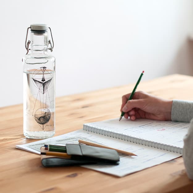 Eine Person, die mit einem Bleistift in ein Notizbuch schreibt. Auf einem Holztisch steht eine dekorative Flasche und ein Etui mit Buntstiften.