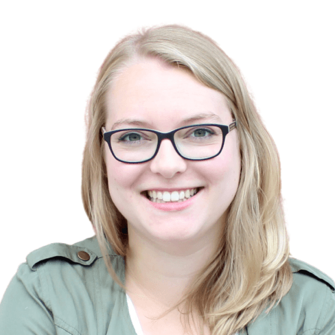 Eine lächelnde Frau mit blonden Haaren und Brille, trägt eine grüne Jacke vor einem weißen Hintergrund.