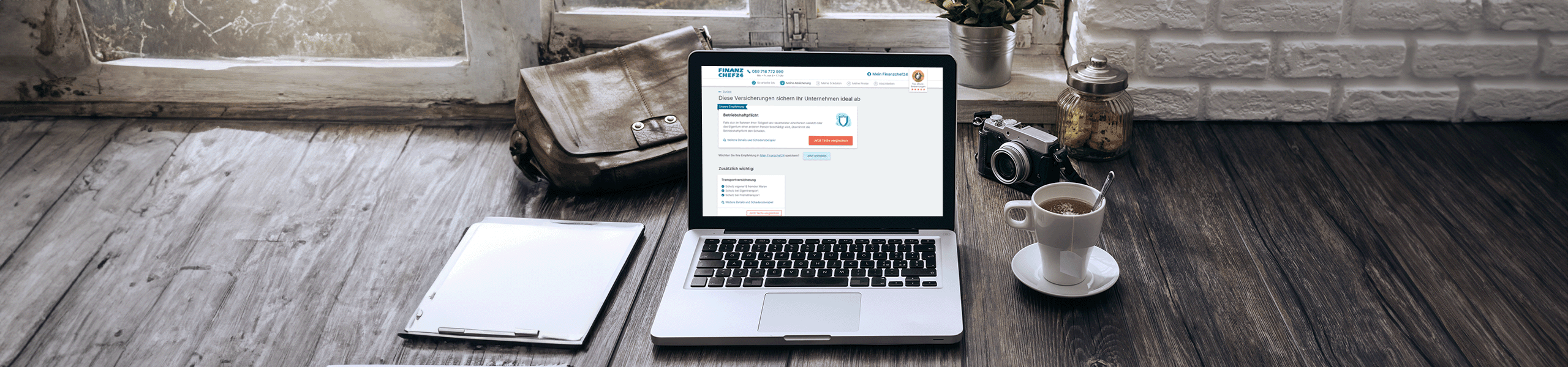Laptop mit Gewerbeversicherungs-Rechner auf einem Holztisch vor einem Fenster mit einer Fotokamera, einer Tasche, einen Kaffee und einem Notizblock in Brauntönen
