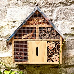 Ein an einer Steinmauer montiertes Insektenhotel aus Holz mit verschiedenen Fächern, die mit Bambusröhren, Holzspänen und Tannenzapfen gefüllt sind.