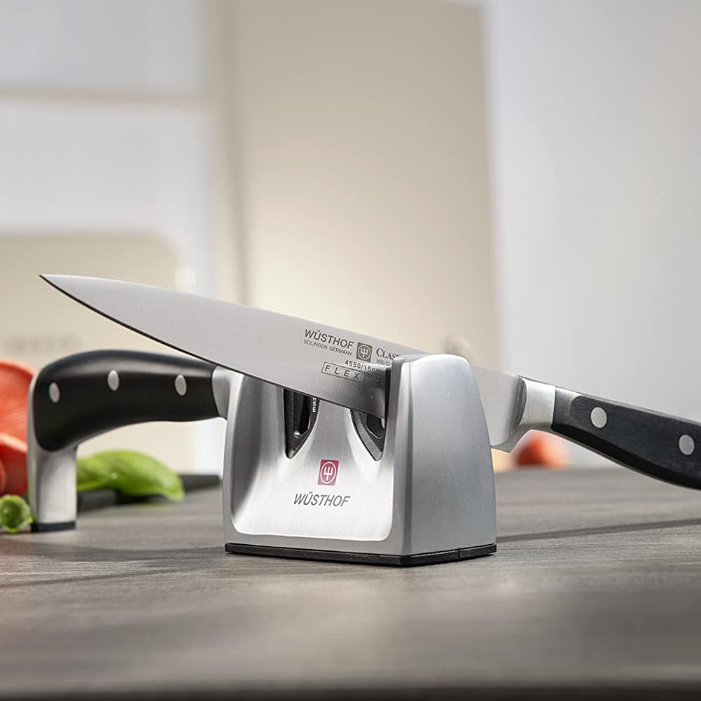 Ein Kochmesser in einem Messerschärfer auf einer Küchentheke, im Hintergrund sind unscharfe Gemüsescheiben zu sehen.