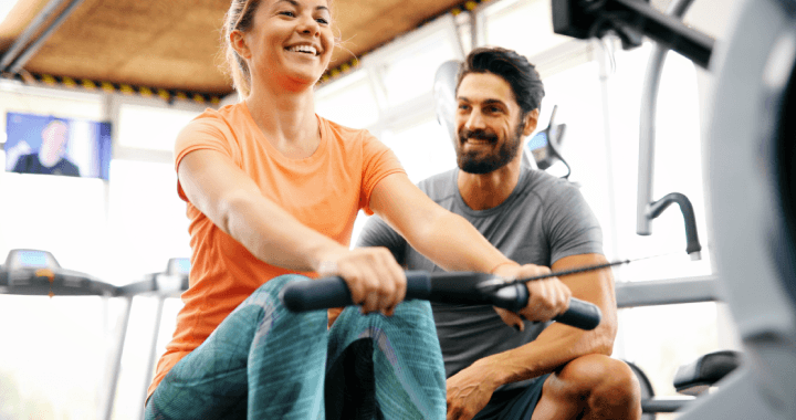 Ein Mann und eine Frau lächeln, während sie in einem Fitnessstudio an Rudergeräten trainieren.