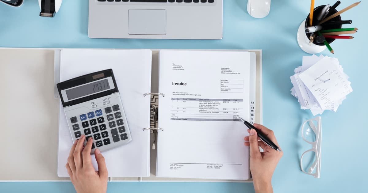 Draufsicht auf einen Schreibtisch mit einer Person, die in einen offenen Rechnungsordner schreibt, umgeben von einem Taschenrechner, einem Laptop, einer Brille und Büromaterial.