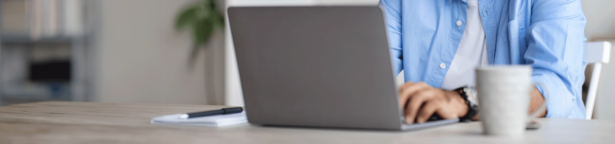 Mann mit blauem Hemd sitzt vor Laptop mit weißem Kaffeebecher.