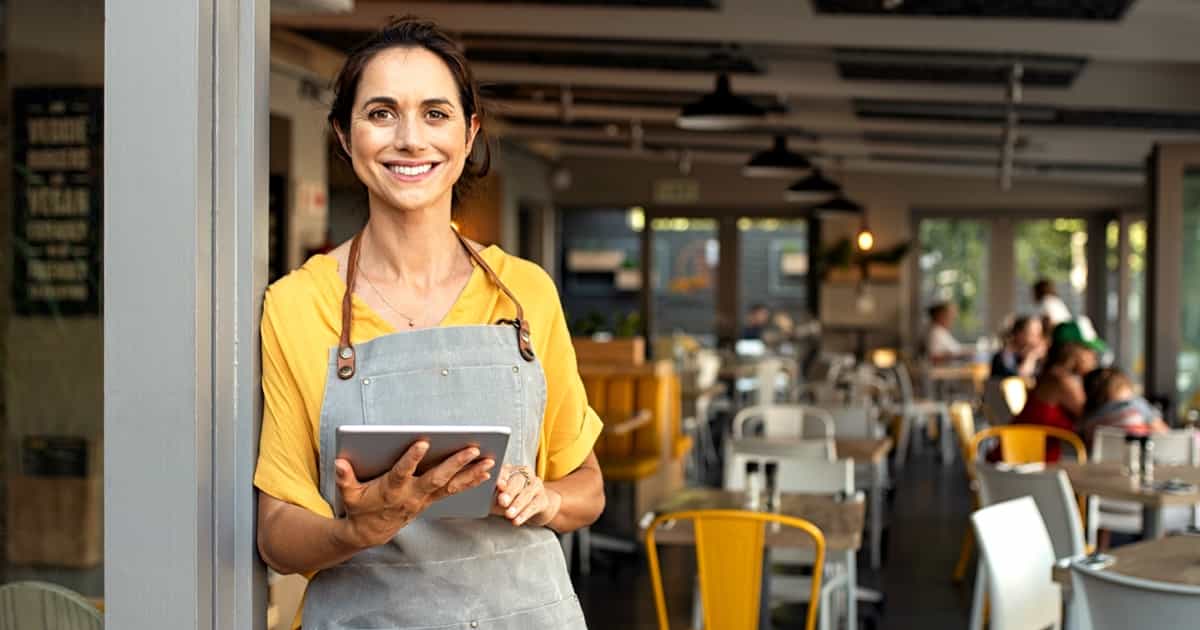 Eine lächelnde Frau in einem gelben Hemd und einer grauen Schürze hält ein Tablet in einem belebten Café mit Gästen im Hintergrund.