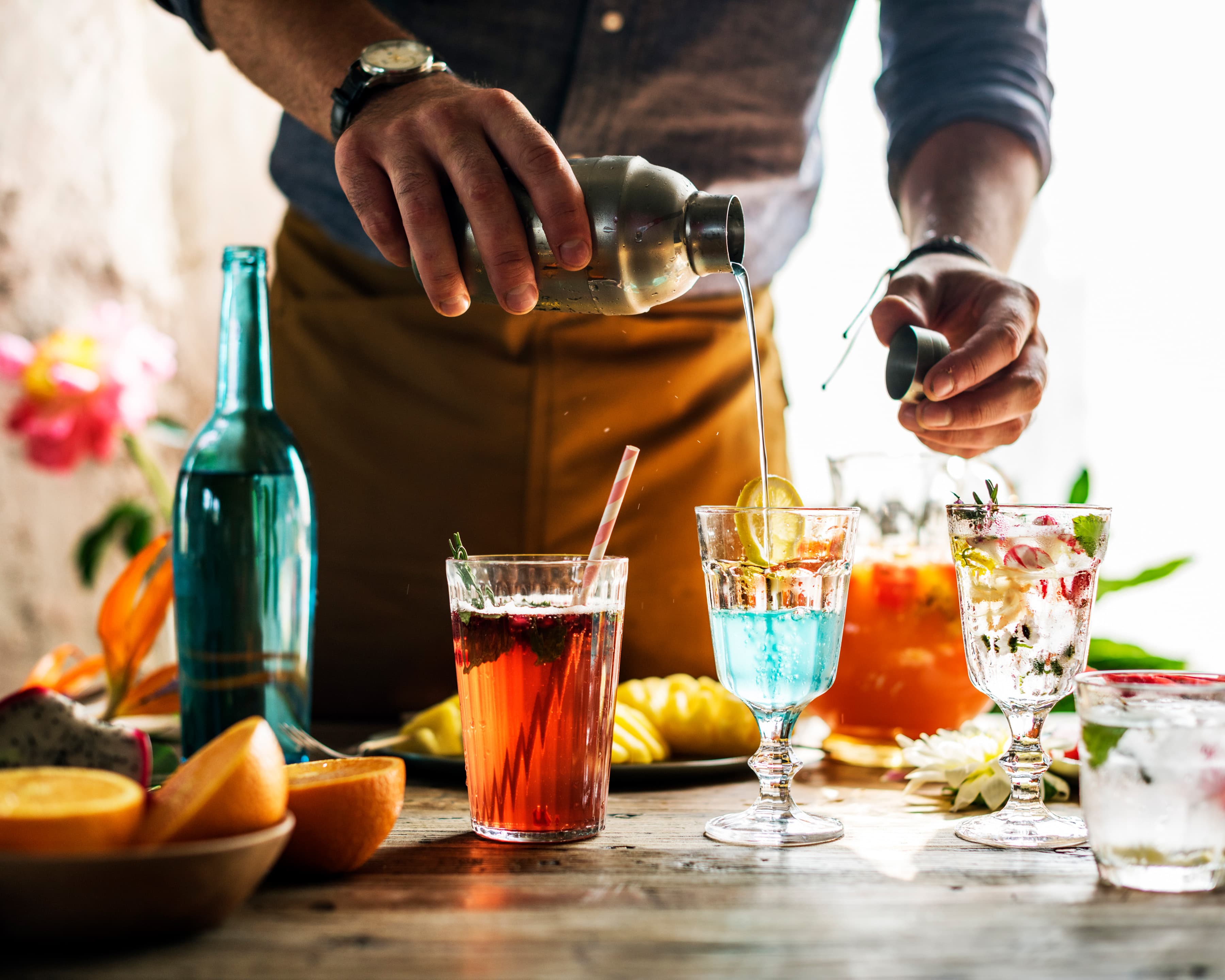 Bartender hält einen Cocktail-Shaker in der Hand und gießt den Cocktail in ein Glas. Um den Cocktail herum befinden sich diverse Materialien zum Cocktailmixen.