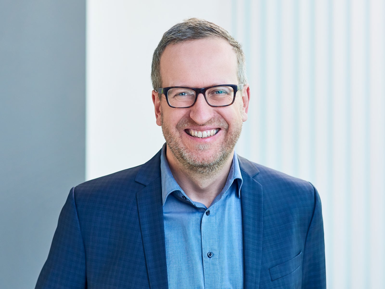 Lächelnder Mann in blauem Anzug und Brille steht vor unscharfem Hintergrund mit vertikalen Linien.